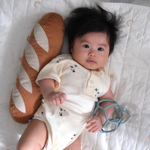 Smiling baby rests on a quilted sheet, cuddling a corduroy baguette-shaped pillow and holding a teal teething toy.
