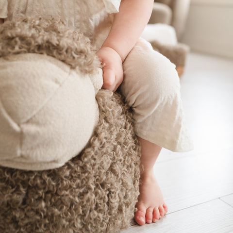 Baby grips a fluffy, tan, sheepskin-textured toy while wearing neutral-toned clothing.
