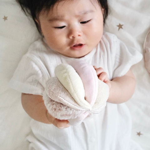 Baby holds a pastel pink, white, and tan flower-shaped soft rattle toy.