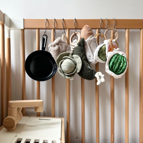 Baby toys, including a toy frying pan, hang from S hooks on a wooden play kitchen.