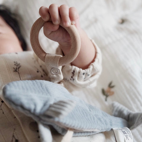 Baby grips a natural wood teething ring attached to a soft, blue plush shark toy.