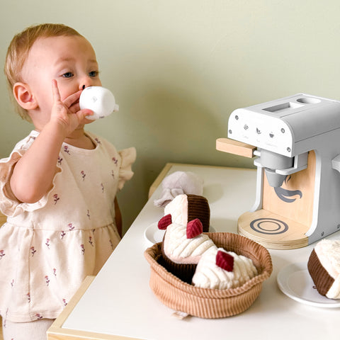 Toddler sips from toy cup at play table with wooden toy coffee maker and plush pastries.