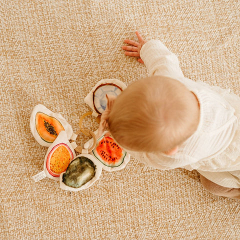 Baby plays with a soft, fabric fruit toy on a beige woven rug.
