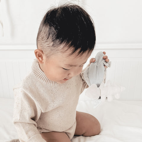 Curious toddler examines a soft, white and grey elephant toy while sitting on a white bed.