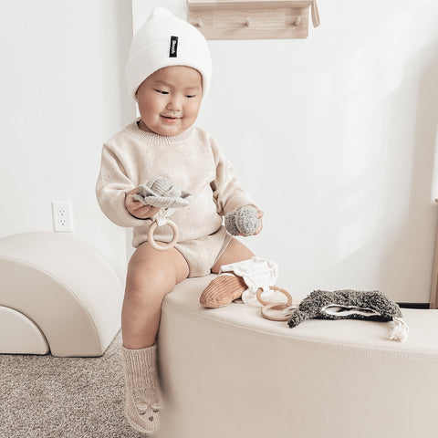 Smiling baby wearing a white beanie explores different textures of baby toys while sitting on a cream-colored bench.