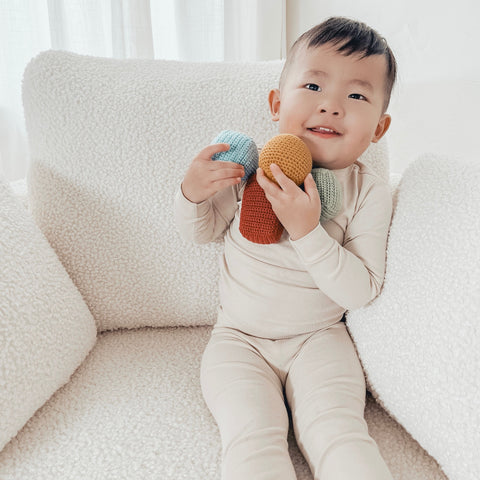 Smiling toddler holds colorful crocheted toys while relaxing on a white sherpa chair.