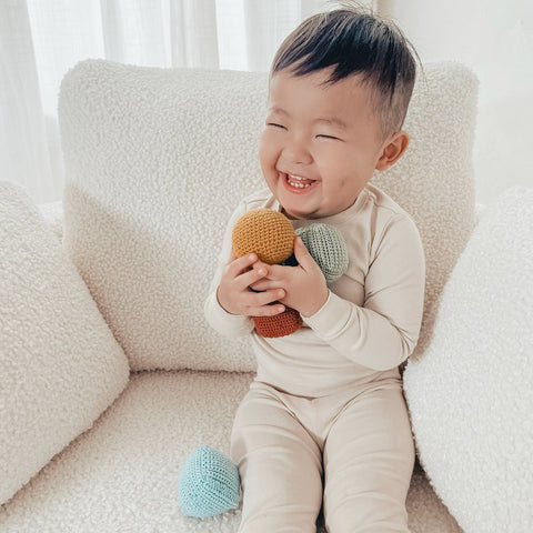 Smiling baby holds colorful crocheted balls while sitting on a cream-colored sherpa chair.