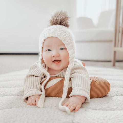 Smiling baby wears a knit pom-pom hat and rests on a brown pillow.
