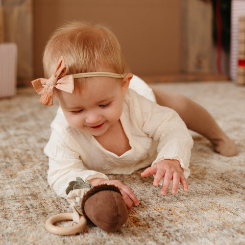 Baby wearing a peach-colored bow reaches for an acorn-shaped baby rattle on a textured rug.