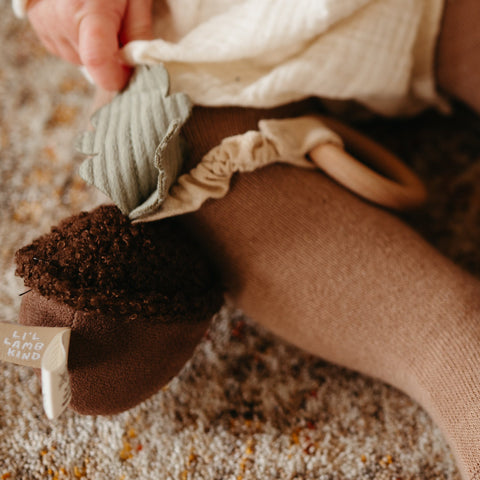 Baby touches a soft, brown Lil' Lamb Kind plush toy with a wooden teething ring.