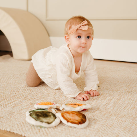 Curious baby wearing a beige bow crawls toward colorful fabric toys.