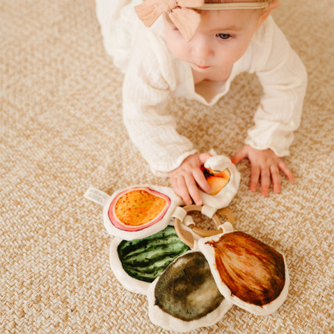 Baby plays with soft, fruit-shaped sensory toys while lying on a beige carpet.