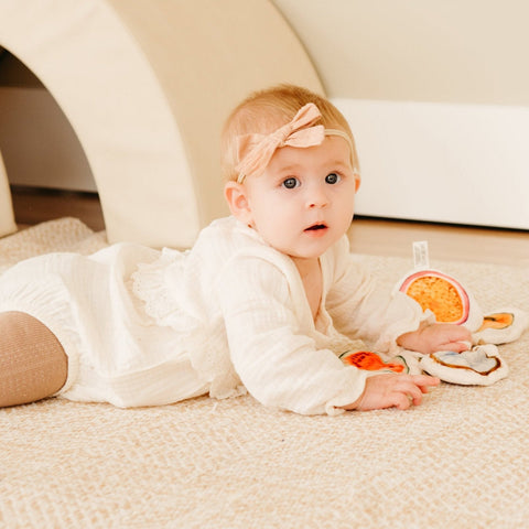 Baby wearing a blush headband plays with soft fruit toys while laying on a beige blanket.