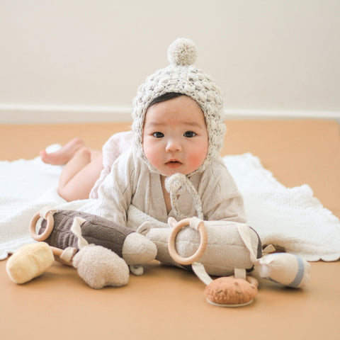 Baby wearing a knitted bonnet explores soft toys while lying on a play mat.