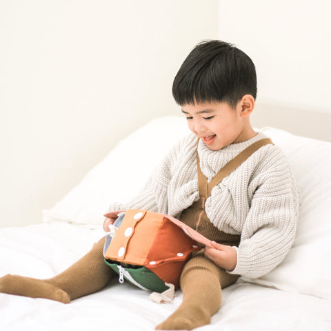 Smiling toddler boy opens a colorful fabric activity cube on a white bed.