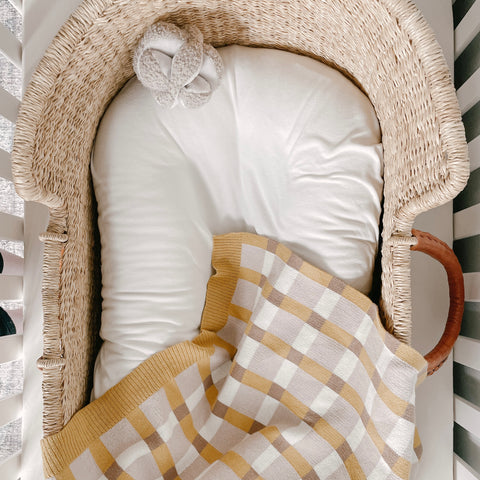An empty woven bassinet sits in a white crib. A yellow and white gingham blanket lays inside, along with a pair of soft baby slippers.