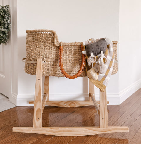 Empty woven bassinet with leather handles rocks gently on wooden base. A soft blanket and plush toys rest inside the bassinet.
