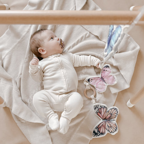 Baby gazes at colorful butterfly toys while lying on a soft blanket under a wooden play gym.