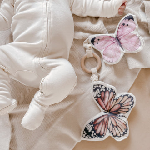 Baby wears soft, cream-colored pajamas while laying beside butterfly-shaped baby toys.