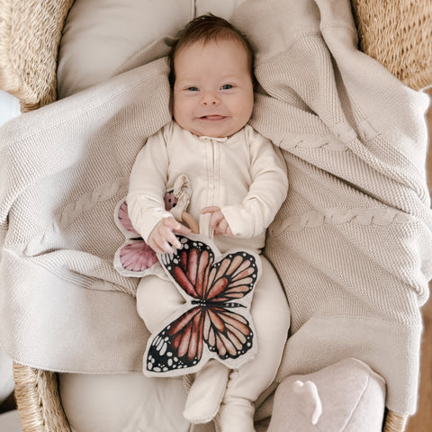 Smiling baby lies in a wicker bassinet on a knit blanket, grasping a butterfly-shaped comfort toy.