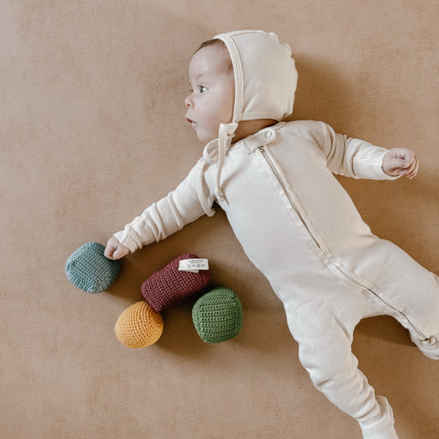 Baby in cream sleeper and matching bonnet lays on tan fabric reaching for colorful crocheted rattle balls.
