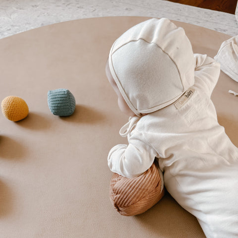 Baby wearing a cream knit baby bonnet plays with crocheted toys on a round play mat.