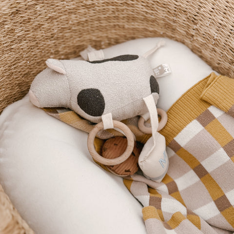 Spotted cow plush toy with wooden teether rings rests in a woven basket alongside a folded baby blanket.