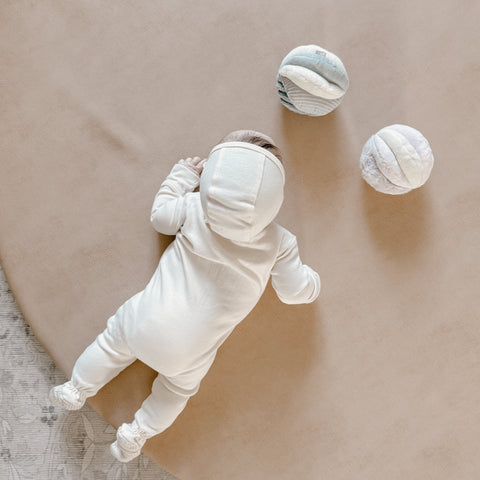 Baby crawls on a soft, tan surface wearing a cream sleeper and plays with textured, plush balls.