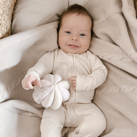 Smiling baby in cream knit onesie holds plush flower toy while lying on a cozy knit blanket.