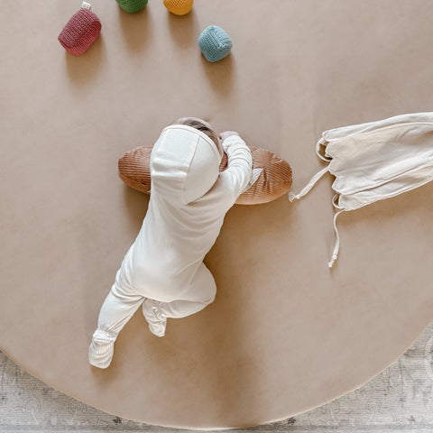 Baby in white plays on a round, beige playmat.