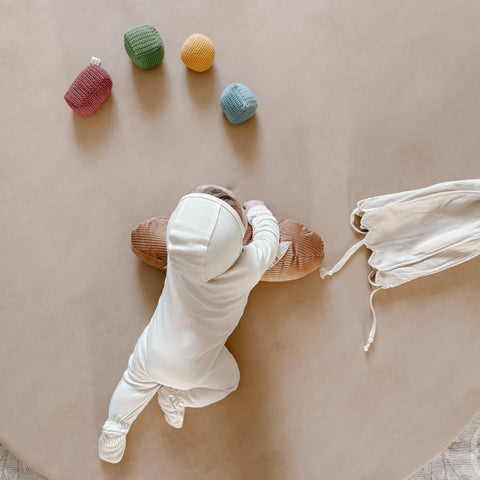 Baby plays on a round, beige play mat. The baby wears a cream onesie and reaches for a brown corduroy bolster pillow. Colorful crocheted balls lay scattered around the baby.