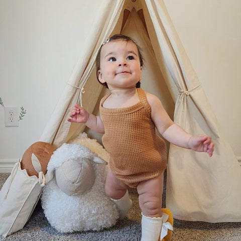 Smiling baby girl stands wearing a waffle-knit romper inside a cream canvas play tent with a fluffy sheep toy beside her.