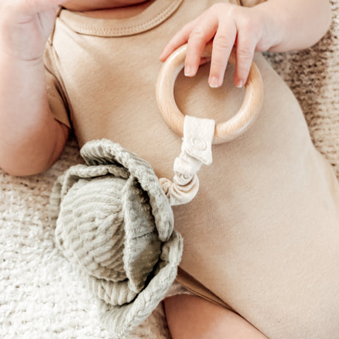 Baby holds a wooden teething ring and soft green toy attached by beige fabric.