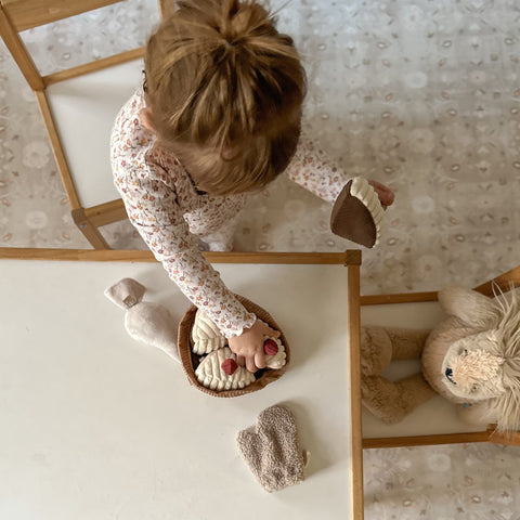 Toddler in floral pajamas reaches for plush toy cupcakes from a fabric basket during playtime.