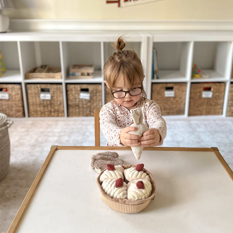Young girl wearing glasses carefully arranges plush pastries on a play table while holding a toy piping bag.