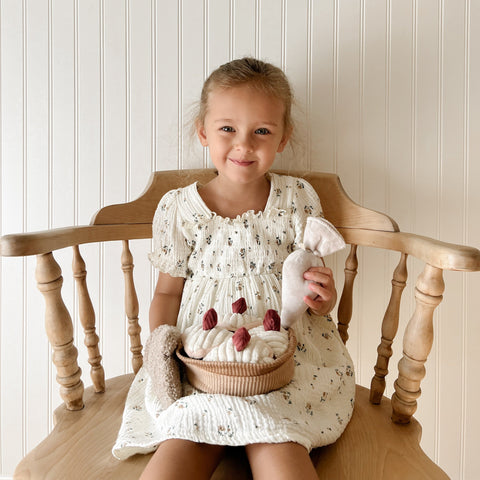 Smiling young girl holds a fabric pie and carrot while sitting in a wooden chair.