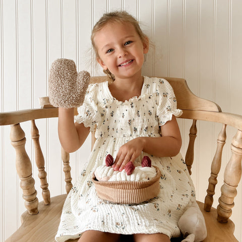 Smiling toddler girl wears fuzzy oven mitt and displays plush toy pie while sitting in a wooden chair.