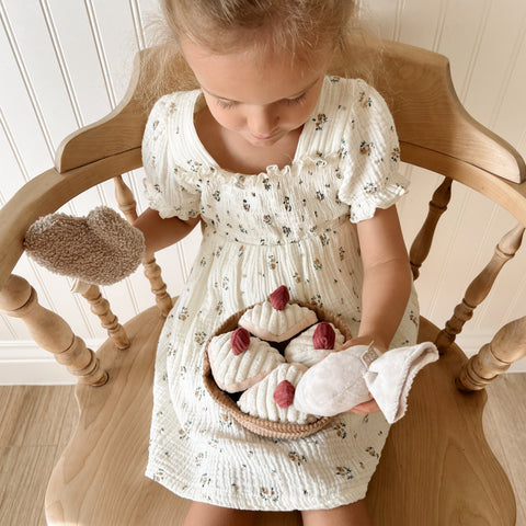 A young child holds a basket of plush toy pastries while sitting on a wooden chair.