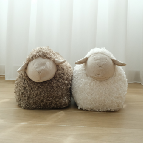 Two fluffy sheep plushes sit on a hardwood floor in front of a window.