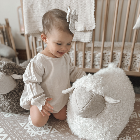 Smiling baby girl reaches for a fluffy white sheep plushie in a neutral-toned nursery.