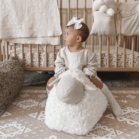 Baby girl with striped bow sits on fluffy sheep toy in nursery with wooden crib.
