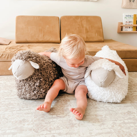 Toddler sits on a geometric rug between a brown and a white plush sheep toy.