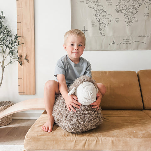 Smiling young boy hugs large stuffed sheep while sitting on a couch.  A world map hangs on the wall behind him.