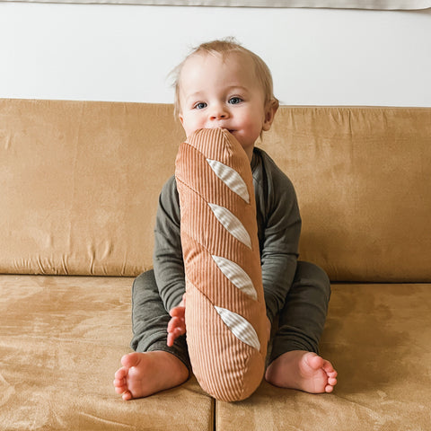 Smiling baby sits on a tan couch and chews on a corduroy baguette-shaped pillow.