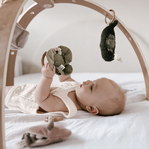 Curious baby explores wooden play gym, reaching for a green corduroy toy.
