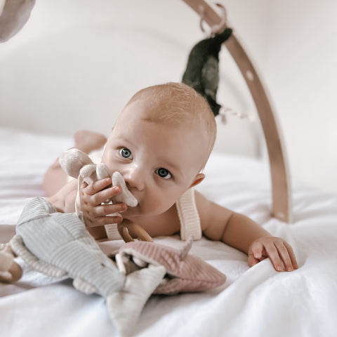 Curious baby chews on a soft toy while lying on a white sheet.