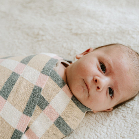 Newborn baby rests on a soft white blanket, swaddled in a pink, tan, and green plaid blanket.