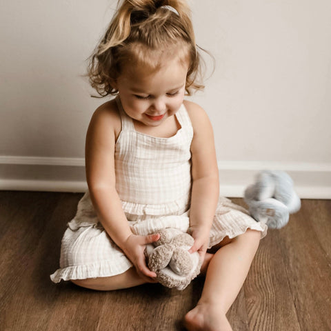 Smiling toddler girl in a gingham dress sits on a hardwood floor playing with a soft, knot-shaped toy.