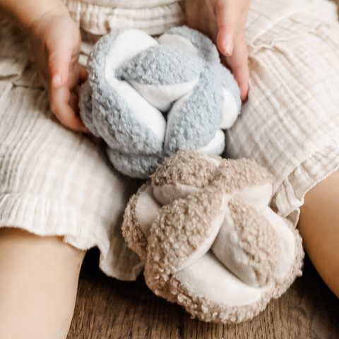 Toddler holds two soft, textured, knotted baby toys in blue and beige.