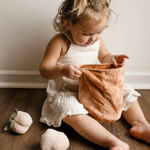Toddler girl in white dress sits on wood floor and peeks inside a soft peach-colored bucket, surrounded by plush peach toys.